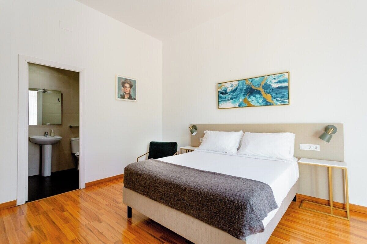 A hotel room with a bed, a bedside table, a lamp, a chair, a sink, and a mirror.