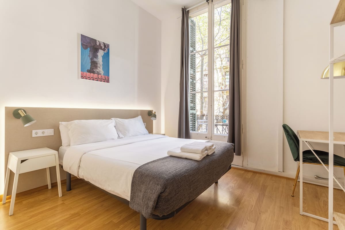 A neatly made bed with a grey blanket, a small bedside table, a desk with a chair, and a window with curtains.