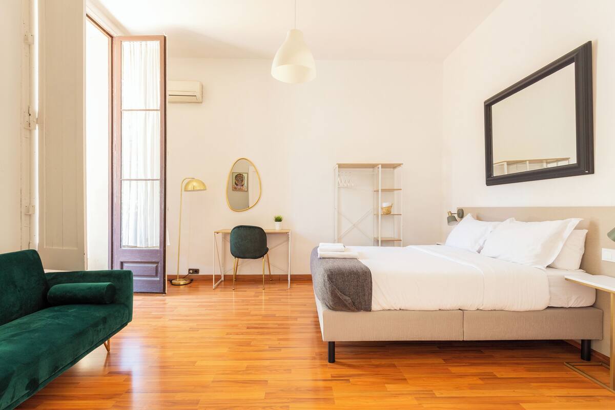 A bedroom with a bed, a desk, a chair, a green sofa, and a large mirror on the wall.