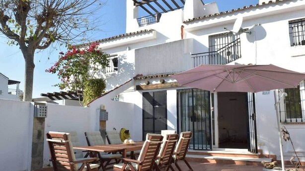 A patio with a wooden table and chairs, a large umbrella, and a white building with balconies. - Casa Jasmine, Modern Villa for 7 M-3 (Marbella)