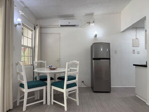 Cozinha compacta com paredes brancas, geladeira de aço inoxidável, mesa redonda com cadeiras e janela com cortinas.