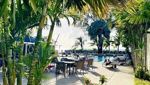 Zona de piscina con sillones reclinables y mesas, rodeada de palmeras y otra vegetación tropical.