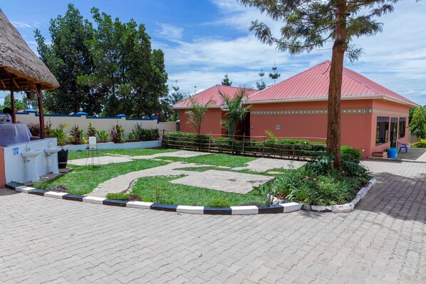 A courtyard with a red-roofed building, a thatched-roof structure, and a paved area with a patterned border. - Mujo Suites Hotel (Sembabule)