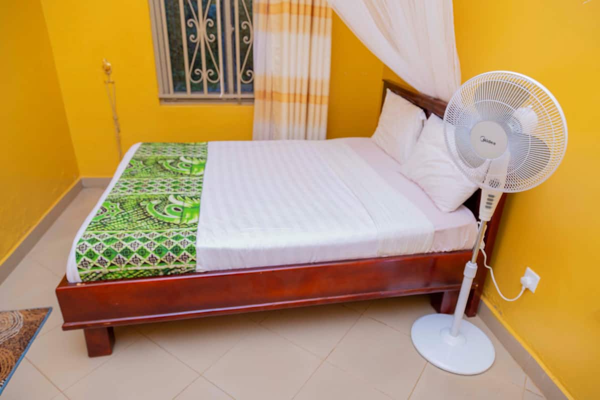 A bed with a white mattress and green patterned blanket, a standing fan, and a window with metal bars and curtains.