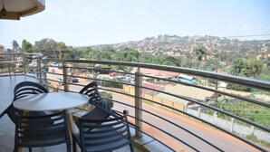 A balcony with a table and chairs, offering a view of a hilly landscape with buildings and a road. - Day 2 Day Prime Hotel (Kampala)