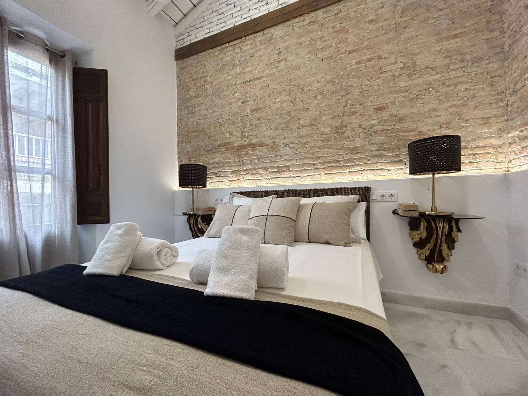 Une chambre moderne avec un mur d’accent en briques, un grand lit avec du linge de lit blanc, deux lampes fixées au mur et une fenêtre avec des rideaux.