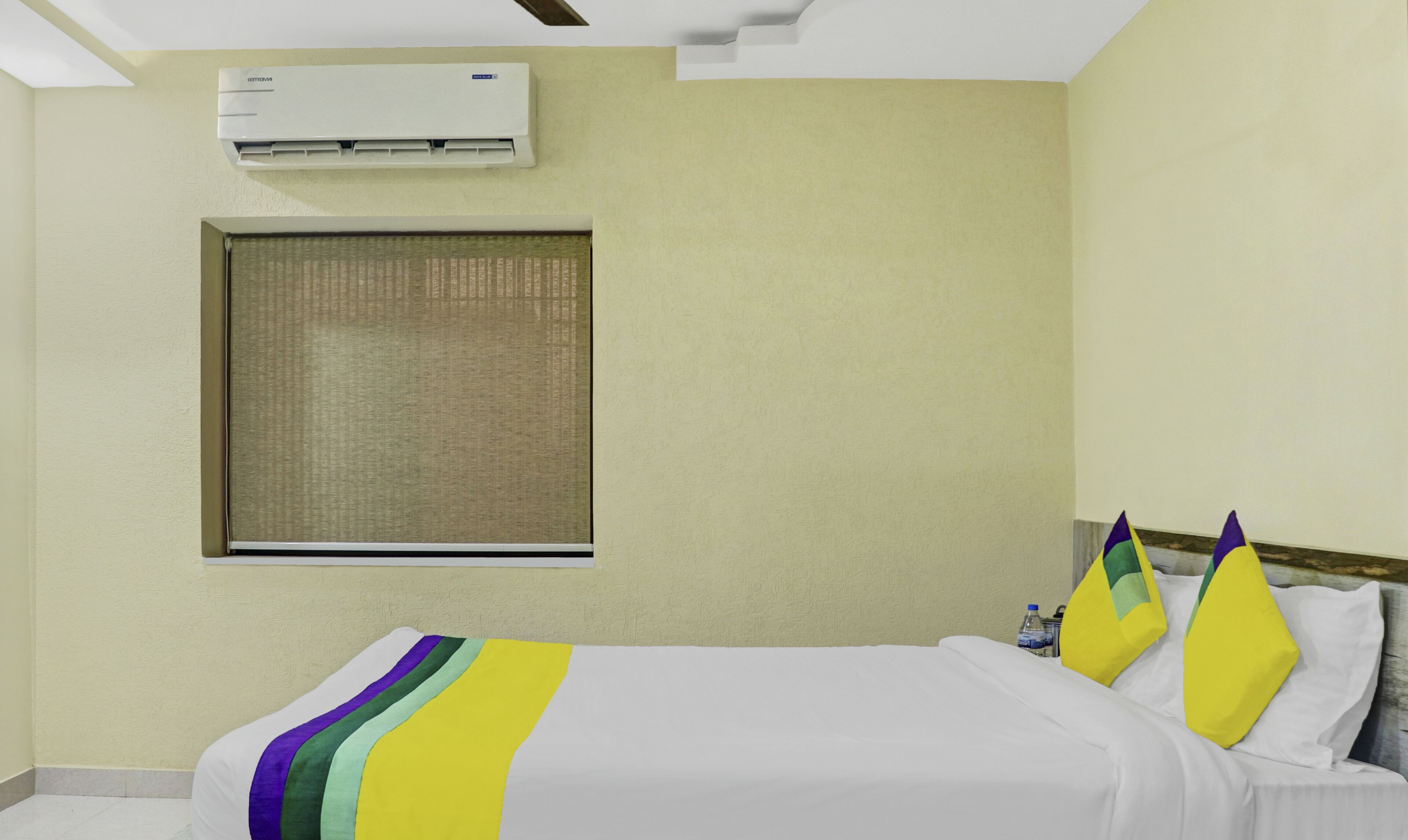 A hotel room with a bed, pillows, a window with a blind, an air conditioning unit, and a water bottle on the nightstand.