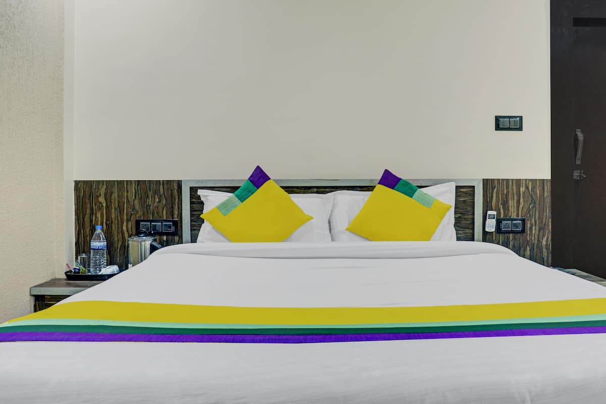 Quarto de hotel com uma cama grande, duas almofadas, uma mesa de cabeceira com uma garrafa de água, um telefone e um interruptor de luz fixado na parede.