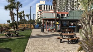 A paved walkway with picnic tables and benches, palm trees, and a building with a sign for a restaurant offering pizza, burgers, fish fry, and fish shanty.