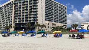 A large hotel with multiple floors and balconies, located on a sandy beach with several beach chairs and umbrellas.