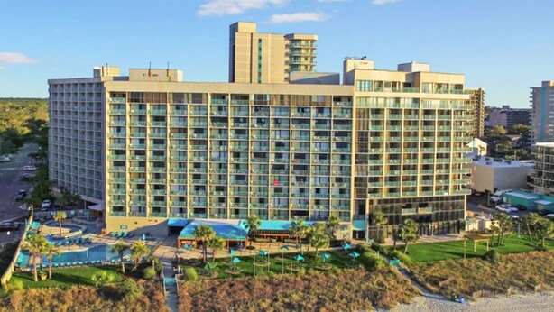 A large hotel with multiple floors, a swimming pool, and a landscaped area with palm trees.