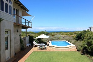 A house with a swimming pool, a patio with a table and chairs, and a view of the ocean. - The Ocean Villa | Mediterranean-Style Beachfront Escape | Pool | Spectacular. (Sandbaai)
