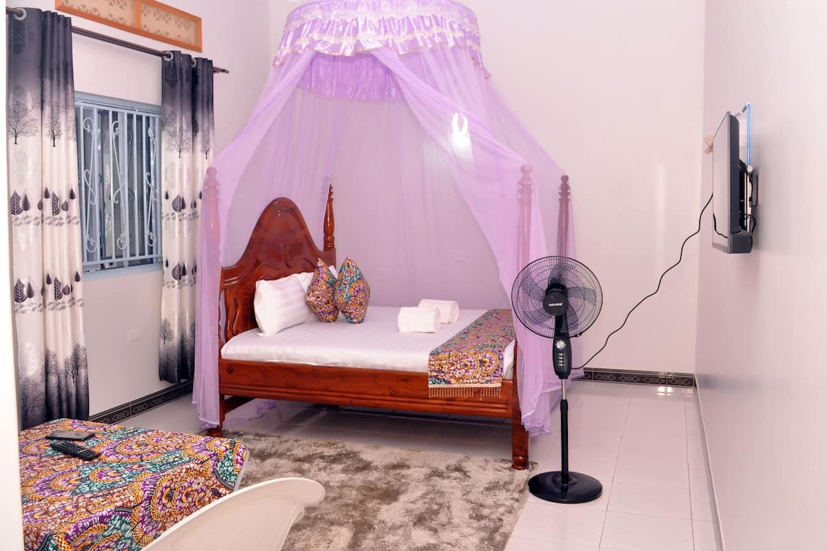 Un dormitorio con una cama con dosel, una mesita de noche, una silla, un ventilador de pie y un televisor montado en la pared.