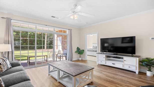 A spacious living room with a large TV, a sofa, a coffee table, and a sliding glass door leading to a patio. - 813 Cypress Bend - Barefoot Resort Rentals (North Myrtle Beach)