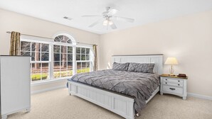 A bedroom with a bed, a nightstand, a lamp, a ceiling fan, and a window with curtains. - 813 Cypress Bend - Barefoot Resort Rentals (North Myrtle Beach)