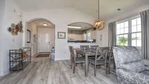 Un espace repas avec une table et des chaises en bois, une cuisine à l’arrière-plan et une vue sur l’extérieur grâce à de grandes fenêtres.