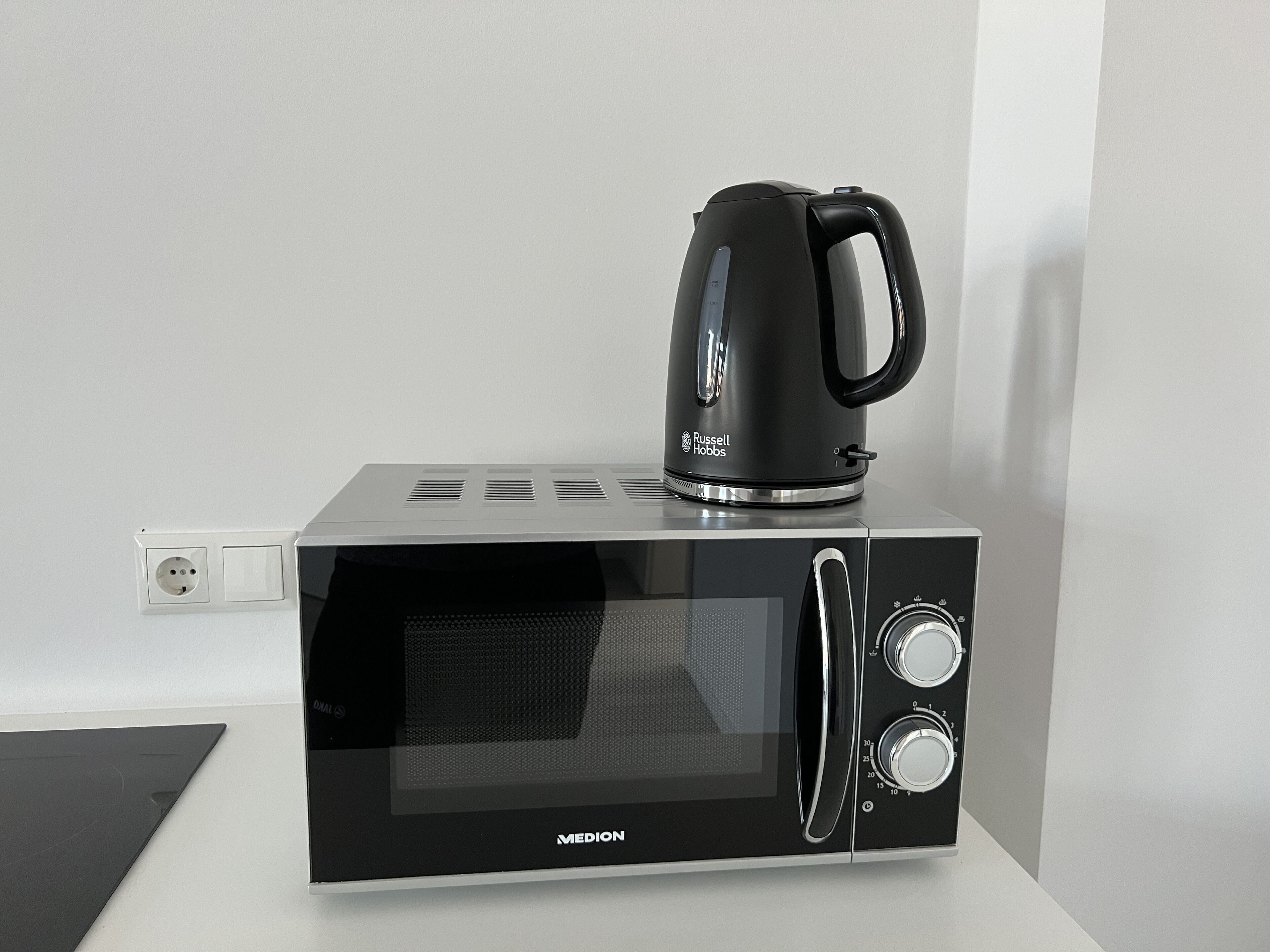 Een zwarte magnetron met een koffiezetapparaat erop, een stopcontact en een bedieningspaneel met twee knoppen.