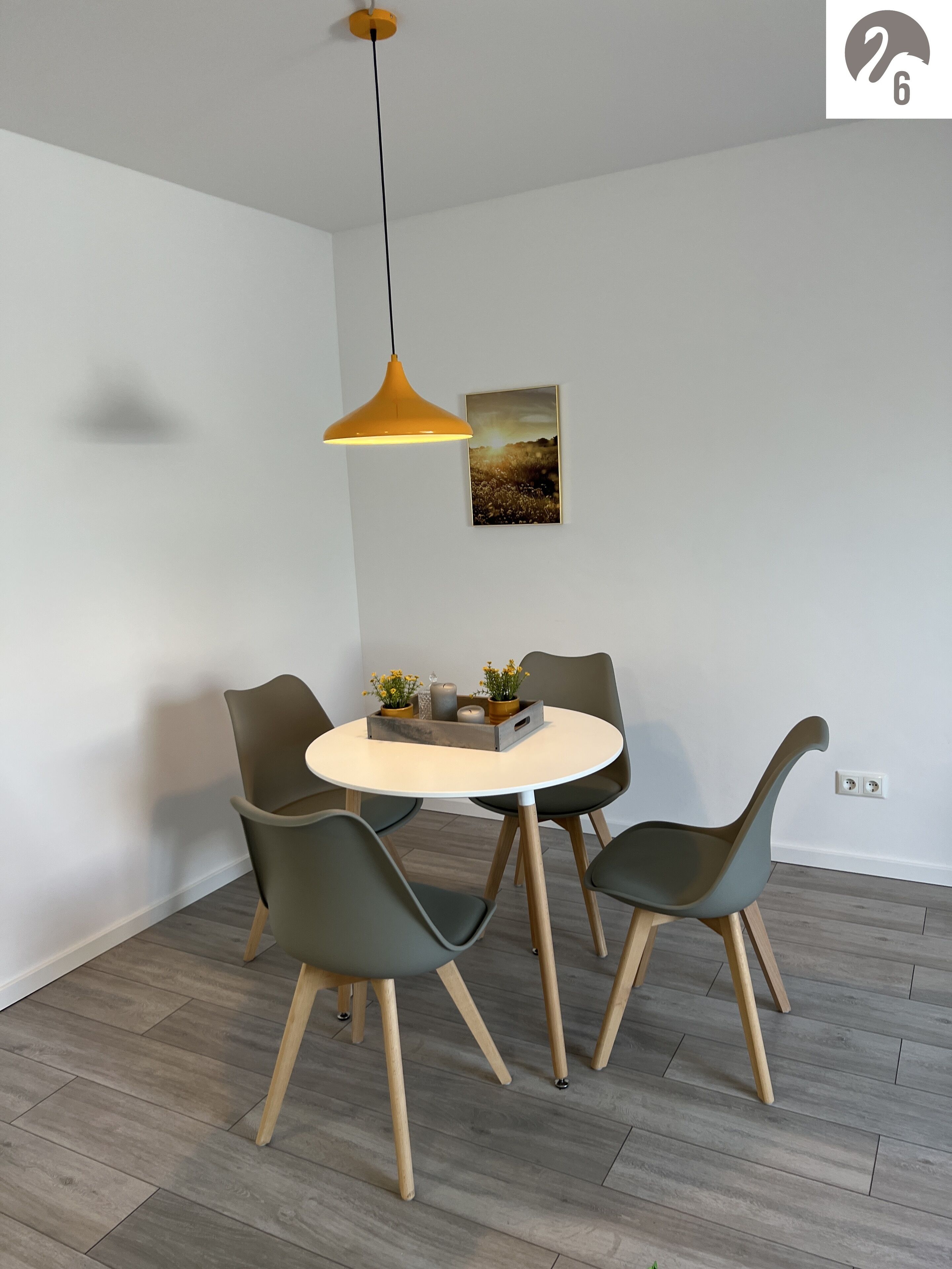 A dining area with a round table, four chairs, a pendant light, and a framed picture on the wall.