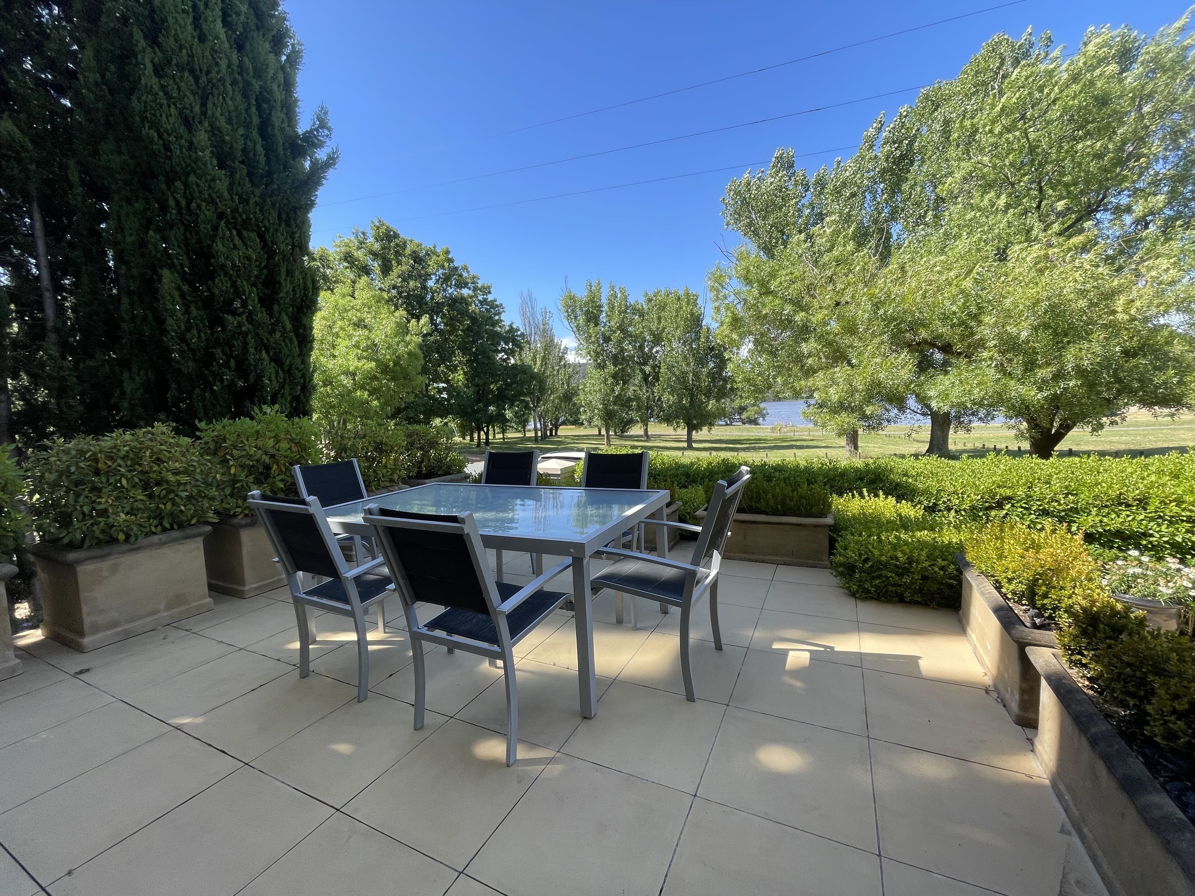 An outdoor patio with a glass table and chairs, surrounded by greenery and trees.