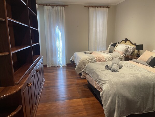 Ein Schlafzimmer mit zwei Betten, Holzboden, einem Kleiderschrank und einem Fenster mit durchsichtigen Vorhängen.