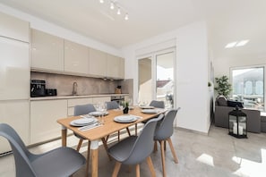 A modern kitchen with a built-in coffee machine, a dining area with a wooden table and grey chairs, and a view of the city through large windows.