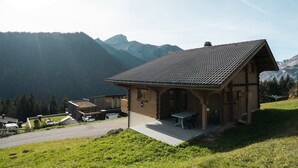 Un chalet doté d’un toit en pente, d’une terrasse avec une table et des chaises, et offrant une vue sur les montagnes en arrière-plan.