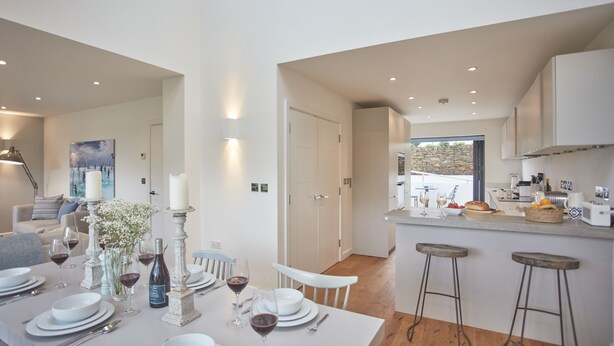 Un comedor moderno con una mesa blanca dispuesta para cuatro, velas y un jarrón de flores. Se ve una cocina con protector de piedra y taburetes de bar al fondo.