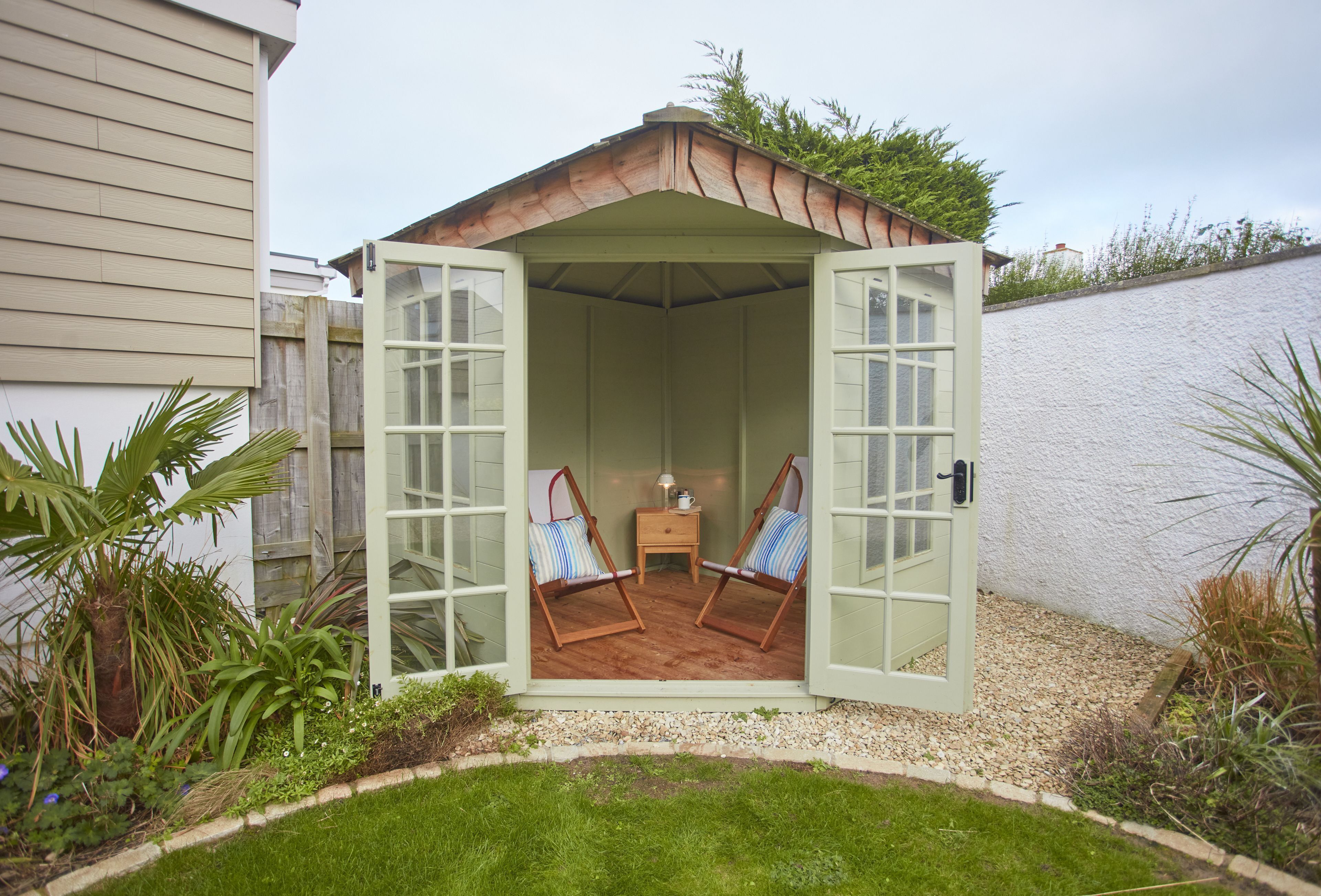 A small, open-sided shed with two cushioned chairs and a side table, surrounded by a garden.