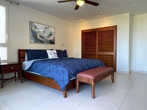 A bedroom with a bed, a nightstand, a bench, a ceiling fan, a wooden wardrobe, and a painting on the wall.