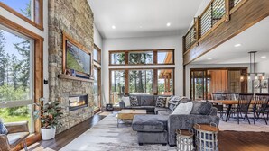 A spacious living room with a stone fireplace, large windows, a high ceiling, and a dining area. - Family-Friendly Luxury | Great Amenities + 1PM Check-In & Check-Out (Cle Elum)