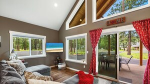 Un salon moderne avec un grand canapé gris, un plaid rouge et un téléviseur à écran plat fixé au mur. Il y a deux grandes fenêtres ornées de rideaux à motifs rouges, ainsi qu’une porte-fenêtre coulissante donnant sur une terrasse extérieure.