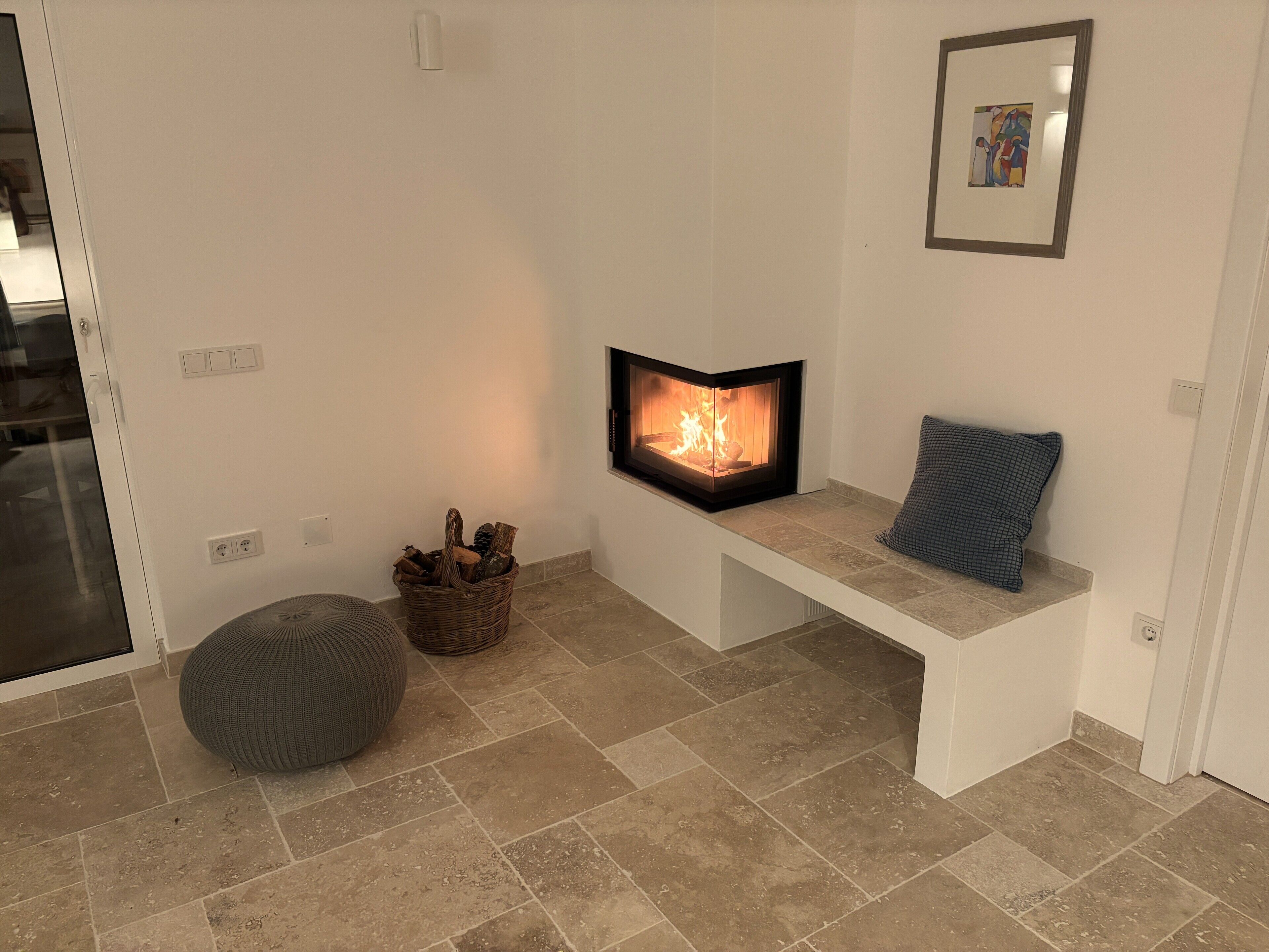 Un salon moderne avec une cheminée, un banc, un panier de bois de chauffage et un pouf gris.