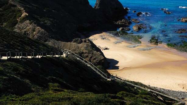 Eine Küstenlandschaft mit Sandstrand, felsigen Klippen und einer Treppe, die zum Ufer hinabführt.