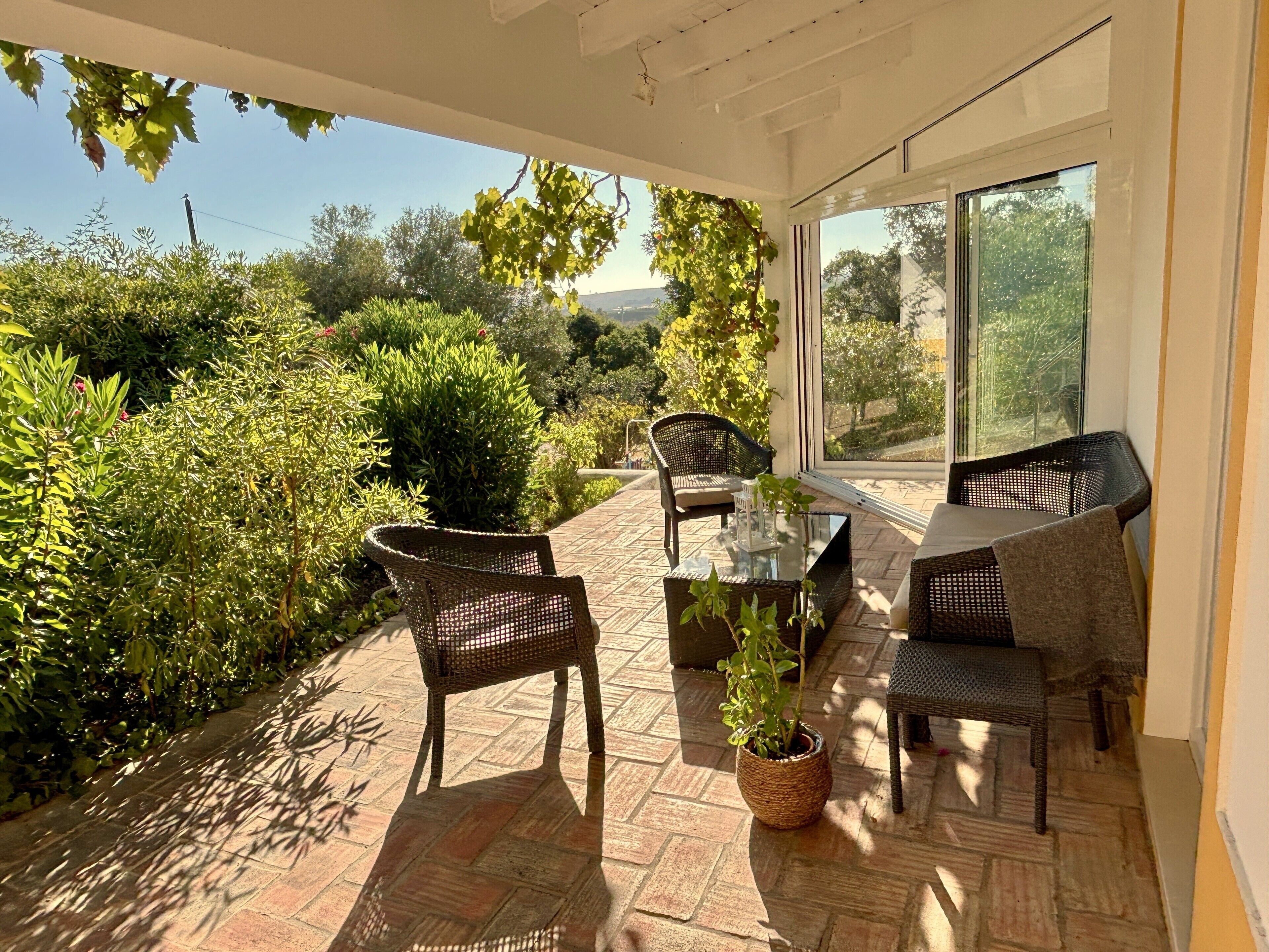 A sunlit patio with wicker furniture, a glass table, and a view of lush greenery and distant hills.