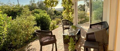A sunlit patio with wicker furniture, a glass table, and a view of lush greenery and distant hills.