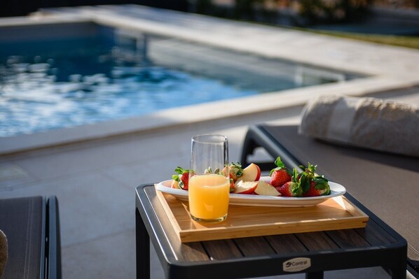 A dining table with a tray of fruit and a glass of orange juice, set on a terrace with a pool in the background.