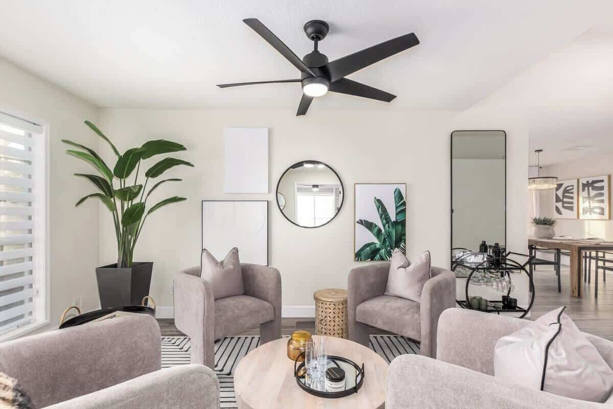 A modern living room with two grey armchairs, a round coffee table, a ceiling fan, a large potted plant, and a dining area in the background.