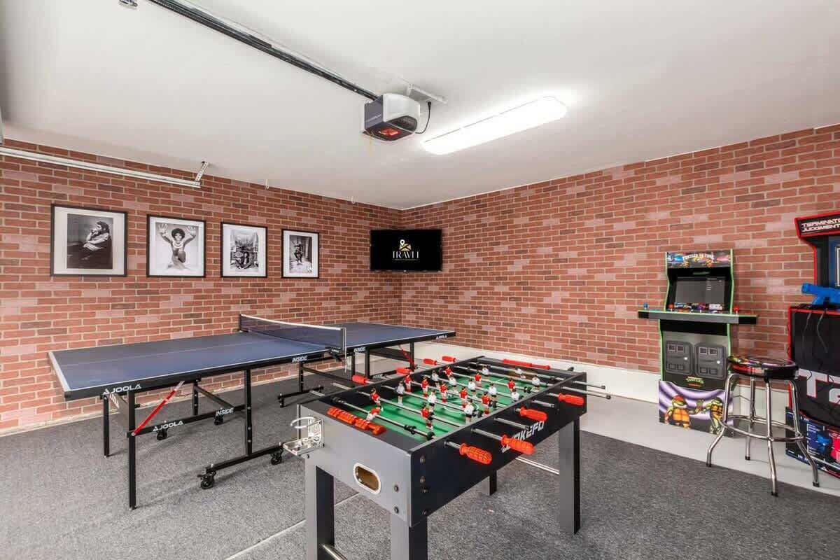 A room with a foosball table, two ping pong tables, and a TV screen.