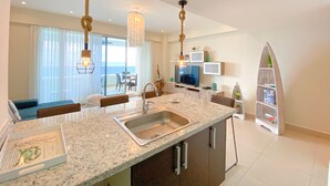 Eine moderne Küche mit einer großen Kochinsel, einer Arbeitsplatte aus Granit und Edelstahlgeräten. Es gibt einen Essbereich mit Tisch und Stühlen sowie einen Blick auf das Meer durch große Fenster.