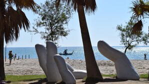 Uma praia com esculturas brancas imponentes, palmeiras e um barco na água.