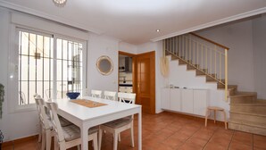 Uma sala de jantar com uma mesa e cadeiras brancas, uma cozinha ao fundo e uma escada de madeira.
