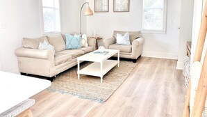 Een zithoek met een beige bank, een witte salontafel en een beige fauteuil. Twee ingekaderde foto's hangen aan de muur, en er staat een vloerlamp.