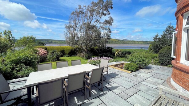 Eine Terrasse im Freien mit Tisch und Stühlen, umgeben von Grünpflanzen und Blick auf einen See.