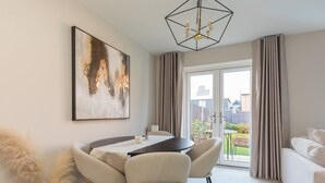 A dining area with a round table, four chairs, a chandelier, a large abstract painting, and a view of a garden through a sliding door.