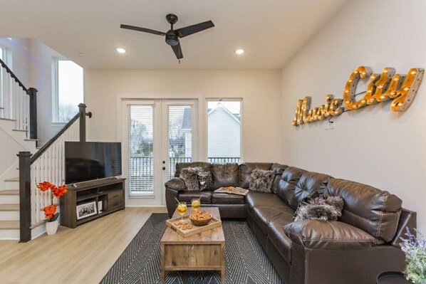 A modern living room with a sectional sofa, a coffee table, and a TV. There is a staircase with a railing and a ceiling fan. The room has a large window and a wall sign that reads "Music City." - ​🎉Celebrate in Style! Game Room, 🎱Pool Table,  💃🏼 Dance Pole! (Nashville)