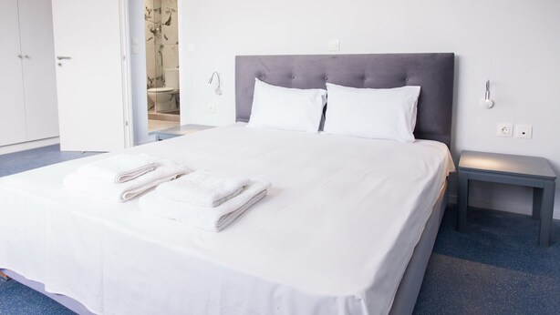 A neatly made bed with white linens and towels, a grey upholstered headboard, a bedside table, and a door leading to a bathroom. - Luxury maisonette in Ilioupoli (Ilioupoli)