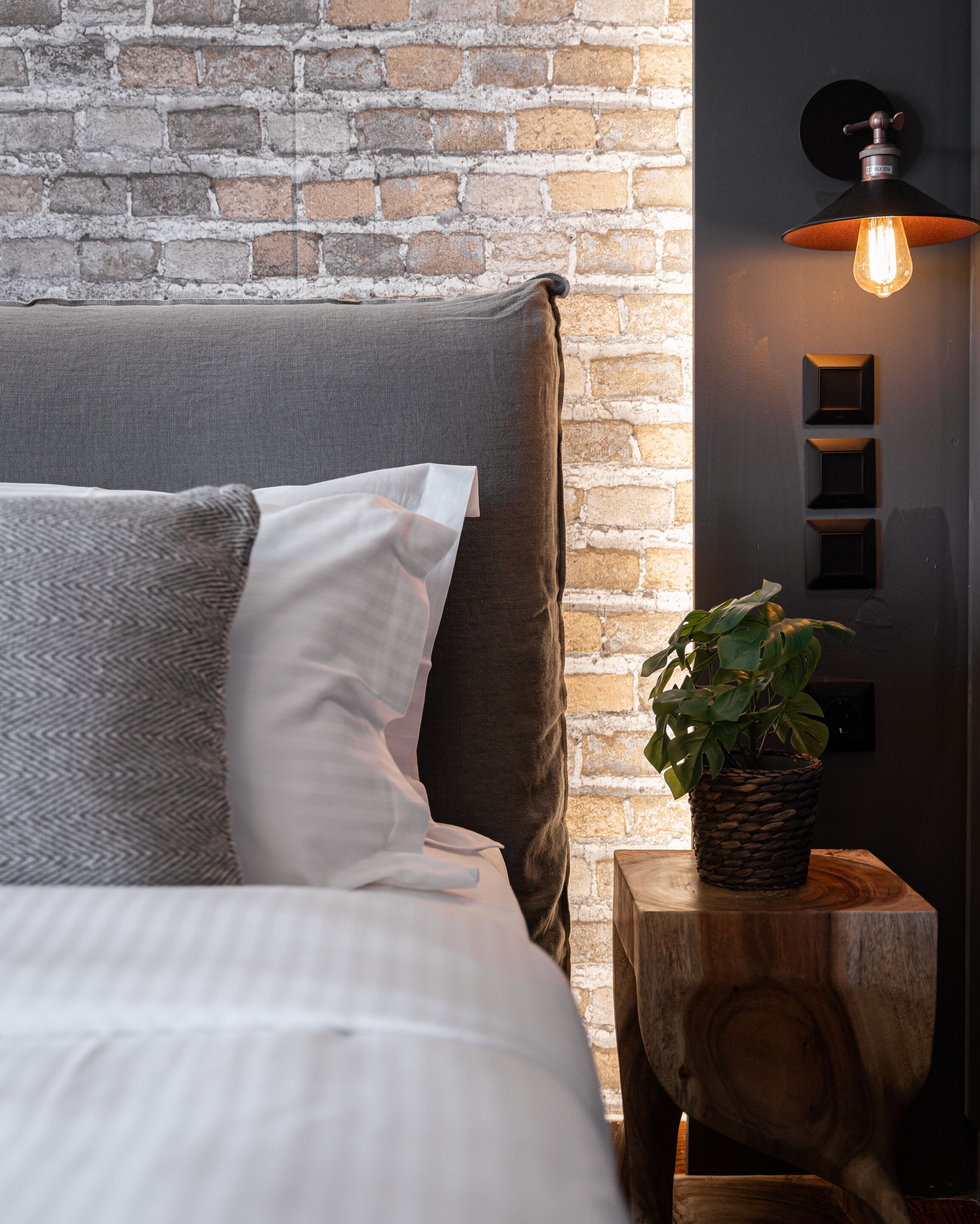 A bed with a grey and white headboard, a potted plant on a wooden nightstand, and a brick wall in the background.