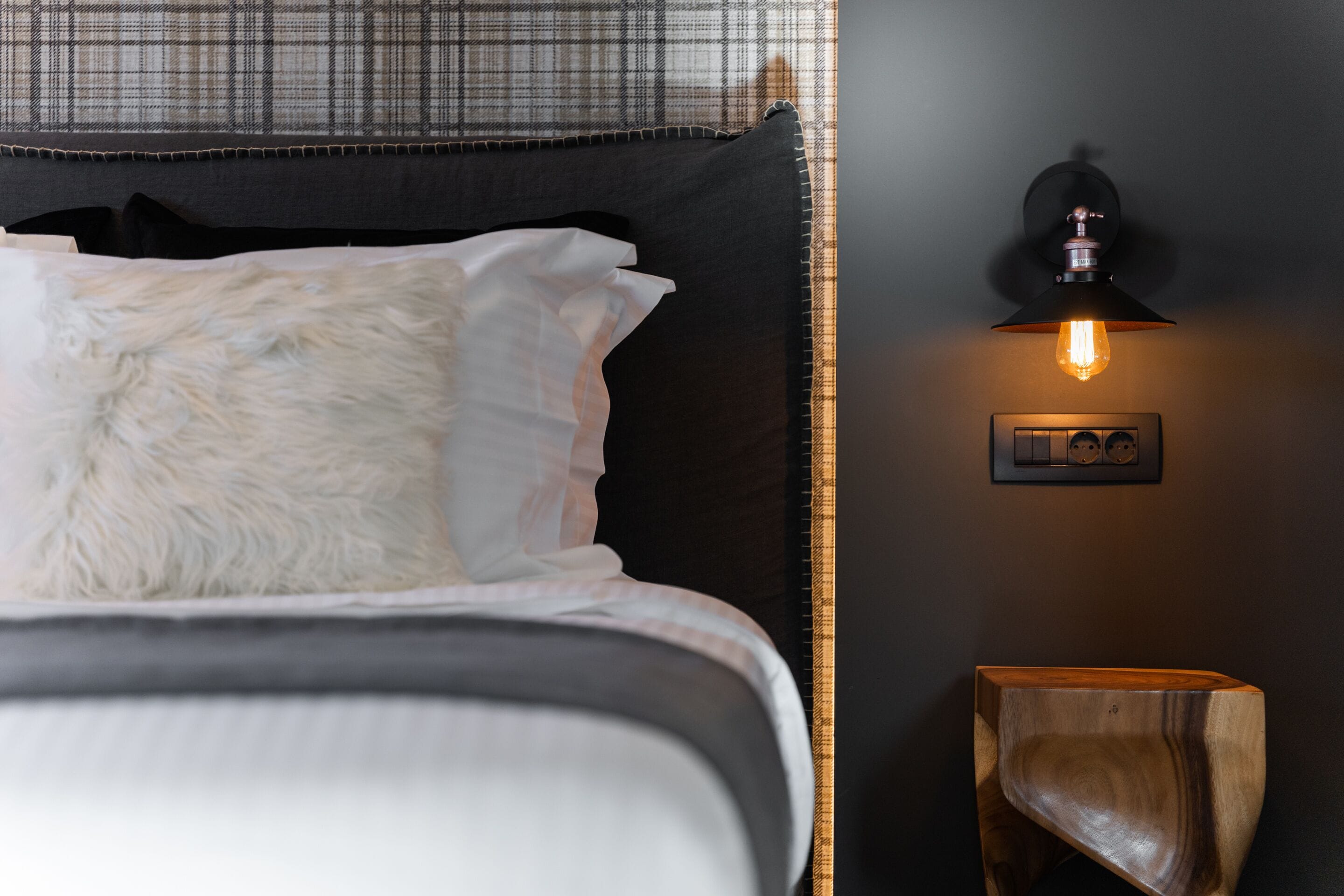 A neatly made bed with white and gray bedding, a fluffy white pillow, and a dark headboard. There is a wall-mounted light fixture with a visible switch and a wooden side table beside the bed.