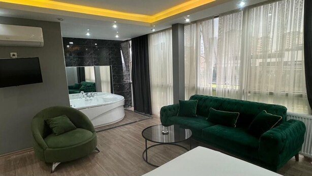 Una habitación de hotel moderna con un sofá verde, una mesita de centro redonda, un televisor de pantalla plana, un ventanal grande con cortinas traslúcidas y una bañera.