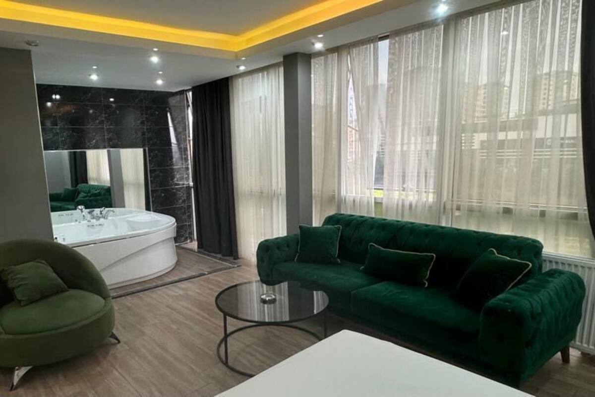 A modern hotel room with a green sofa, a round coffee table, a flat-screen TV, a large window with sheer curtains, and a bathtub.